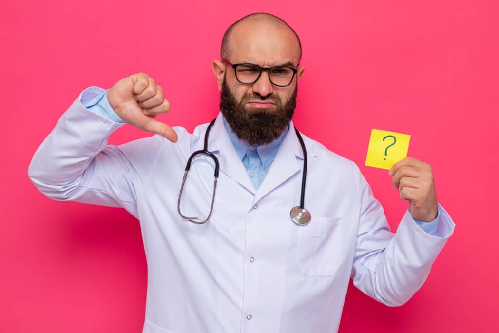 bearded man doctor in white coat with stethoscope around neck wearing glasses holding reminder paper with question mark showing thumbs down indicating questions