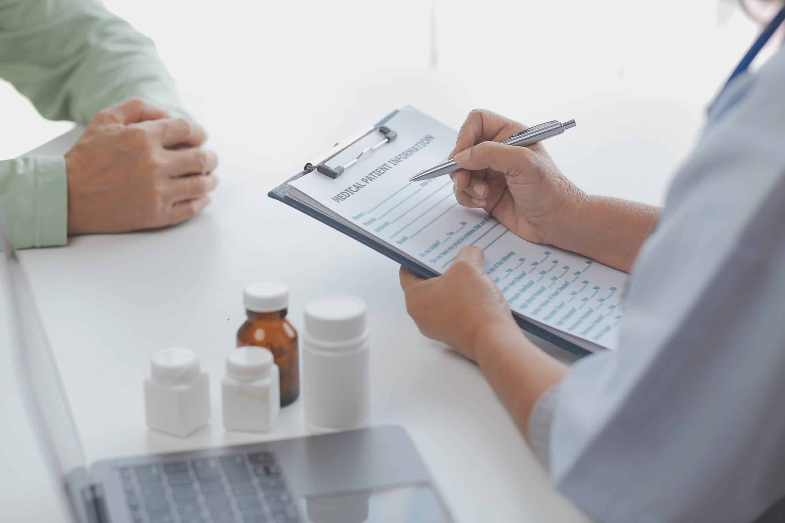 A clinician reviews a patient information form on a clipboard, with medicine bottles on the desk.